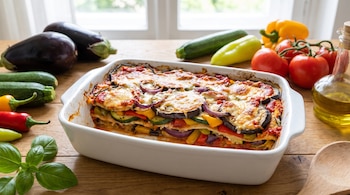 Lasaña en fuente rectangular con capas de berenjena, zucchini, morrones y cebolla, queso gratinado, salsa de tomate, rodeada de vegetales frescos y aceite.