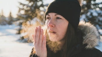Primer plano de mujer con gorro negro y chaqueta de invierno exhalando vapor visiblemente. El sol brilla sobre la nieve y los árboles en el fondo.