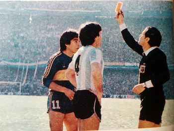 Árbitro con tarjeta amarilla, jugador de Argentina número 10, jugador de Boca Juniors número 11. Campo de fútbol con público en estadio