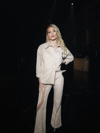 Una mujer rubia con un conjunto de camisa y pantalón beige posa de pie en un estudio con fondo oscuro