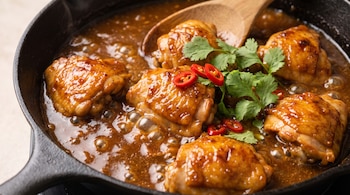 Sartén de hierro fundido con piezas de pollo doradas cubiertas de salsa marrón, decoradas con hojas de cilantro y rodajas de chile rojo.