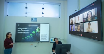Una mujer de pie y un hombre sentado en un aula. Hay dos pantallas grandes, una muestra una presentación y la otra una videollamada con seis personas