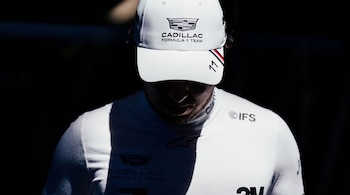 Un hombre con gorra y prenda blancas con el logotipo del equipo Cadillac de Fórmula 1 e insignias de patrocinadores mira hacia abajo.