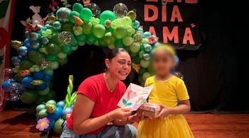 Pamela Franco, con una blusa roja, sonríe emocionada mientras mira un dibujo. A su lado, una niña con vestido amarillo sostiene un regalo frente a un arco de globos.
