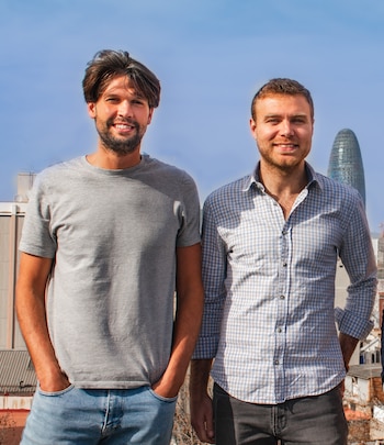 Primer plano de dos hombres. Uno lleva camiseta gris y vaqueros, el otro camisa a cuadros. Fondo urbano con edificio cilíndrico y cielo azul