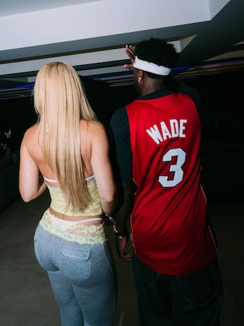 Dos personas de espaldas: una mujer rubia con top amarillo de encaje y pantalón gris; un hombre con camiseta roja de baloncesto "WADE 3" y jeans