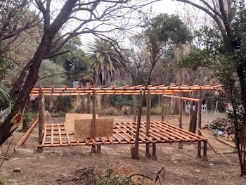 Estructura de madera en construcción con postes y vigas, cajas de madera en el centro, terreno de tierra, árboles, palmera y vegetación
