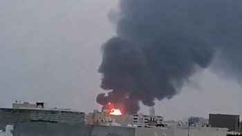 Un gran incendio con llamas brillantes y una enorme columna de humo negro denso se eleva sobre una zona edificada bajo un cielo gris y cubierto