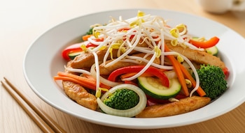 Plato hondo blanco con chop suey de pollo, tiras doradas, zanahoria, morrón rojo, cebolla, zucchini, brócoli y brotes de soja. Palitos chinos a un costado en mesa de madera clara.