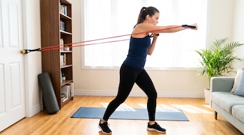 Mujer con ropa deportiva haciendo ejercicio con bandas de resistencia rojas ancladas a una puerta, en una sala con estantería, esterilla, planta y sofá.