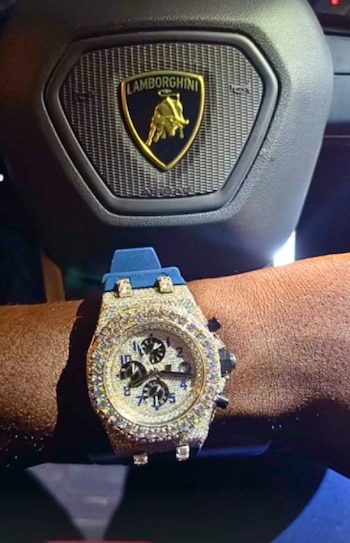 Una muñeca con un reloj de pulsera cubierto de pedrería y correa azul frente al volante de un automóvil con el emblema de Lamborghini
