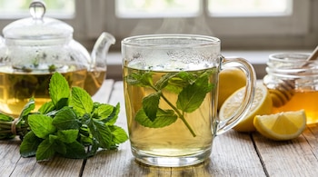 Taza de infusión caliente con hojas de menta, tetera de vidrio, manojo de hierbabuena, limones, tarro de miel con palillo en mesa de madera.