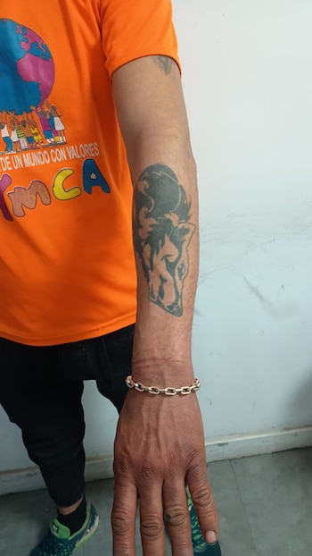 Un hombre viste una camiseta naranja y muestra un tatuaje oscuro de un animal en el antebrazo junto a una pulsera dorada