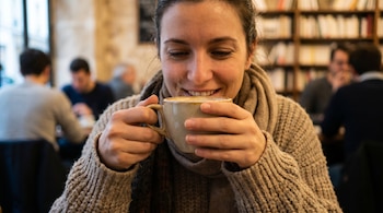 Primer plano de una mujer sonriente con suéter y bufanda, sosteniendo una taza de café humeante en sus manos, con personas y estanterías borrosas al fondo.
