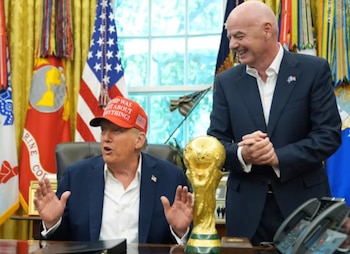 Donald Trump con una gorra roja y Gianni Infantino sonriendo junto al trofeo dorado de la Copa Mundial de la FIFA en una oficina con banderas