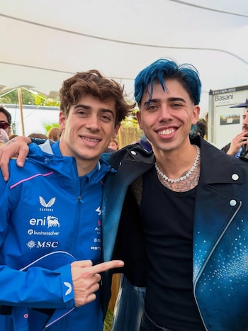 Franco Colapinto, en chaqueta azul con logos deportivos, sonríe junto a Luck Ra, con cabello azul, chaqueta de cuero negra y azul, y collar brillante