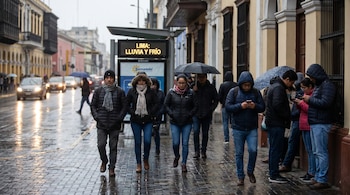 Personas abrigadas caminan por una calle mojada en Lima bajo un cielo gris. Un cartel digital muestra "LIMA: LLUVIA Y FRÍO". Algunos revisan sus teléfonos.