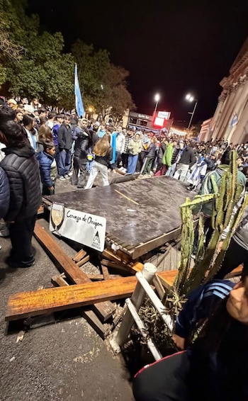 Grupo de personas sobre una pérgola de madera con un cartel de "Gustito", ondeando banderas de Argentina, con una multitud debajo y un edificio al fondo