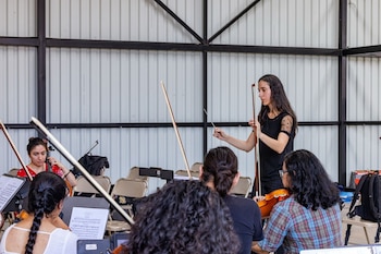 Vista de una directora de orquesta, de cabello oscuro, que dirige a músicos sentados con instrumentos de cuerda y partituras en un salón con paredes de metal corrugado
