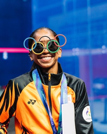 Una mujer con chaqueta deportiva naranja y negra sonríe y usa gafas con la forma de los cinco aros olímpicos