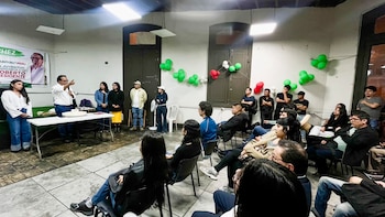 Un hombre habla en un evento con una audiencia de personas sentadas y de pie. Globos verdes, blancos y rojos decoran la pared. Un cartel de campaña es visible detrás