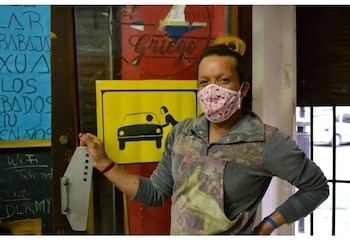 Persona con mascarilla estampada y ropa gris sostiene un medidor metálico gris. Al fondo, un cartel amarillo con un dibujo de un coche y una persona