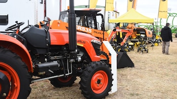 Primer plano de un tractor naranja con ruedas negras y detalles metálicos en una exhibición al aire libre. Se ven otras máquinas al fondo