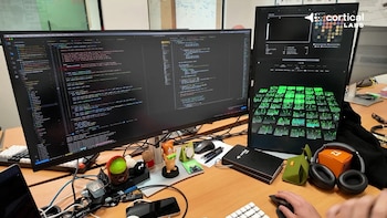 Escritorio con dos monitores de computadora mostrando código de programación y gráficos de datos neurales, ratones, teclados y otros objetos personales