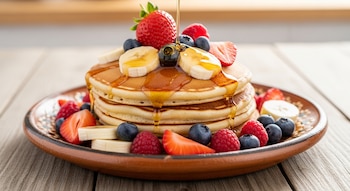 Una pila de pancakes dorados con fresas, arándanos, frambuesas y rodajas de plátano es rociada con miel dorada, servida en un plato rústico.