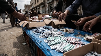 Fotografía de un puesto callejero improvisado con un mantel azul, mostrando blísteres de medicamentos de colores apilados, billetes, monedas y manos manipulándolos.