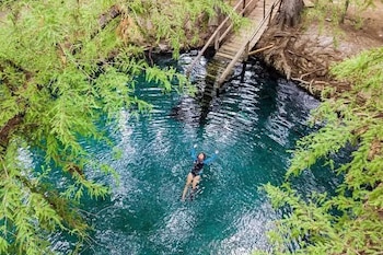 Vista aérea de una persona flotando boca arriba en agua turquesa rodeada de árboles frondosos y una pasarela de madera en la orilla