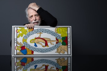 Hombre mayor con barba blanca, sombrero fedora negro y abrigo oscuro. Manos levantadas hacia adelante, palmas abiertas, sobre un fondo gris