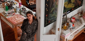 Una mujer con camisa de estampado animal print se apoya en un marco de puerta blanco. Mesas llenas de pinturas, pinceles, lienzos y materiales de arte