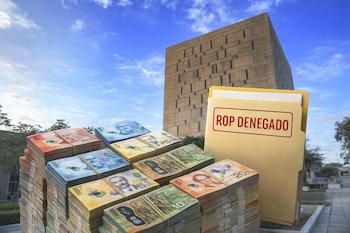 Fajos de billetes de colones de Costa Rica, una carpeta beige con "ROP DENEGADO" en rojo, y un edificio de concreto bajo un cielo con nubes.