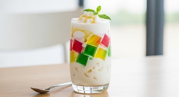 Un vaso alto con un frappé blanco cremoso lleno de cubos de gelatina roja, verde y amarilla, adornado con menta, crema y caramelo sobre una mesa de madera con una cuchara.