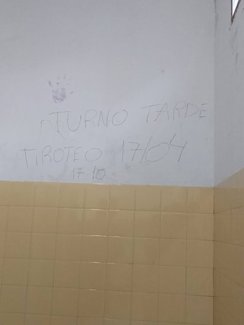 Primer plano de una pared blanca con un mensaje manuscrito en grafito que dice 'TURNO TARDE', 'TIROTEO 17/04' y '17:10'. Debajo hay azulejos marrones