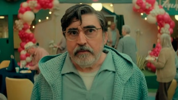 Primer plano del actor Alfred Molina, hombre con barba gris y gafas, vistiendo polo y cárdigan azul claro, mirando al frente. Fondo con globos y personas