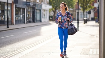 Una mujer sonriente de unos 40 años con ropa deportiva vibrante, leggings azules y zapatillas, camina por una acera con una taza de café y una bolsa de gimnasio.