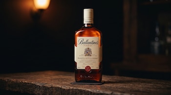 Una botella de whisky Ballantine's Finest sobre una superficie de madera rugosa frente a un fondo oscuro.