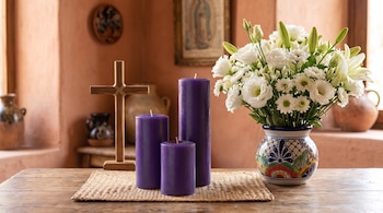 Tres velas cilíndricas moradas, una cruz de madera y un jarrón con flores blancas colocados sobre un tapete tejido en una mesa de madera.