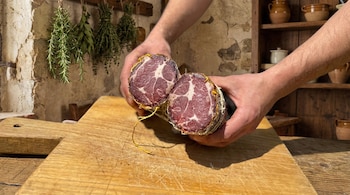 Dos manos sostienen un embutido curado cortado por la mitad, mostrando su interior marmoleado, sobre una tabla de madera. Al fondo, hierbas secas y cerámica.