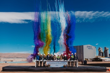 Directivos con cascos blancos en un escenario al aire libre frente a humo de colores y un edificio con el logo de Posco Argentina