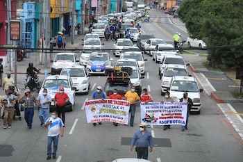 Calle con personas en protesta portando dos pancartas blancas. Numerosos taxis blancos, motocicletas y edificios a los lados. Algunas personas usan mascarillas