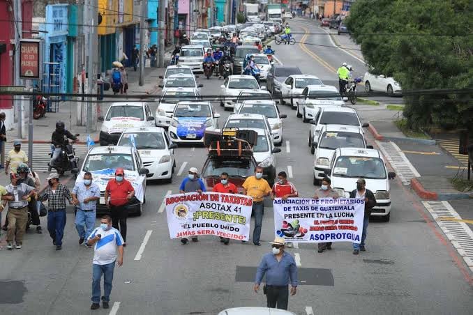 Un grupo de taxistas guatemaltecos se manifiesta en una calle de la ciudad, portando pancartas y acompañados por vehículos de taxi. (Redes Sociales)