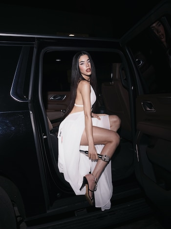 Una mujer con vestido blanco y zapatos de tacón sostiene un micrófono junto a la puerta abierta de un automóvil negro