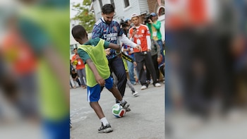 Zohran Mamdani, vestido con una camiseta de fútbol, juega con un balón junto a un niño que lleva un peto fluorescente en una calle