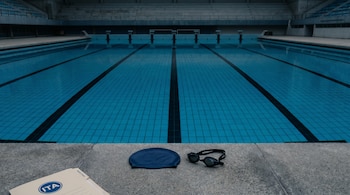 Piscina olímpica vacía de agua azul con líneas de carril, vista desde el borde. Una gorra azul, antiparras negras y un documento con el logo ITA están en el primer plano.