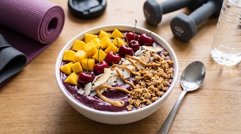 Bowl de açaí con mango, cerezas, coco, mantequilla de maní y granola. Junto a una cuchara, esterilla de yoga morada, mancuernas y botella de agua transparente.