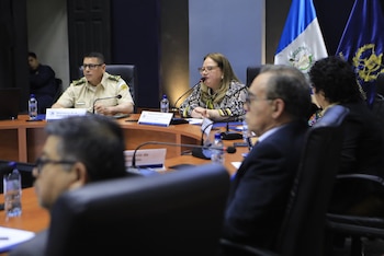 Una mujer con gafas y blusa estampada habla por un micrófono en una mesa redonda junto a un hombre en uniforme militar y otras personas; banderas de Guatemala al fondo
