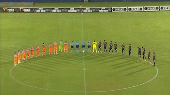 Jugadores con uniformes de color naranja y negro junto a cuatro árbitros alineados en el círculo central de una cancha de fútbol.